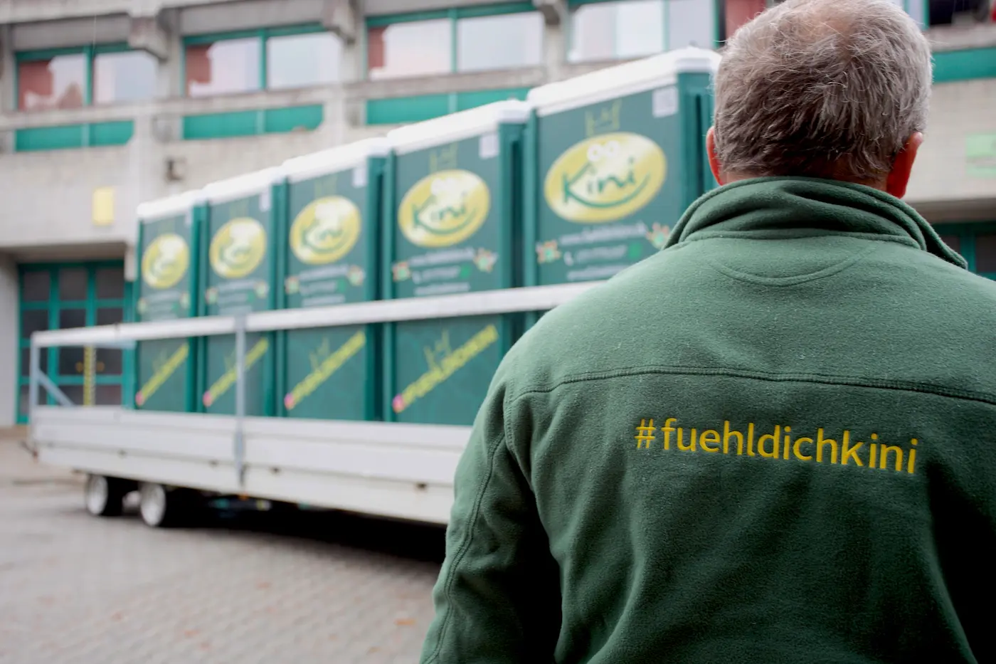 fuehldichkini-heisl-service-mitarbeiter-von-hinten-mit-grünen-sweater-logo-vor-transport-anhänger-mit-fünf-mobile-toilette-vor-betonbau-münchen-für-baustelle-die-eine-baustellentoilette-mieten-dixie-klo-mieten